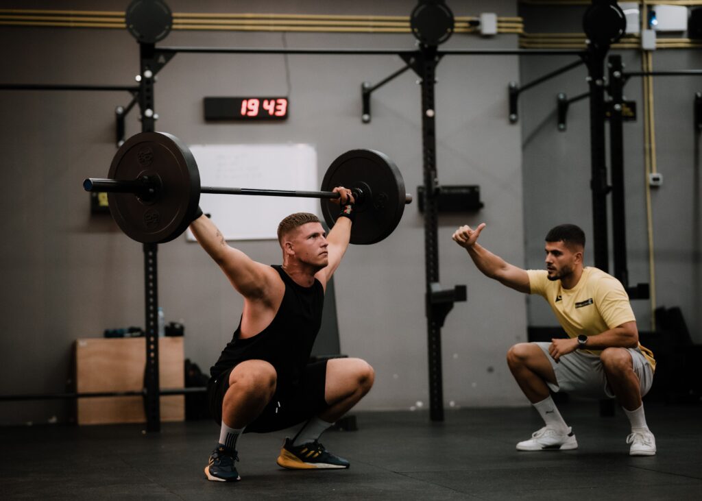 Fisioterapia Deportiva A muscular man performing a weightlifting squat with a coach guiding him indoors.