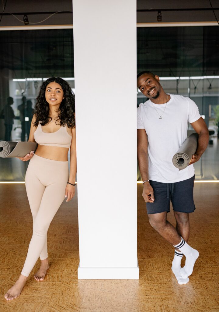 Pilates Reformer A black man and woman standing indoors with yoga mats, ready for a fitness class.
